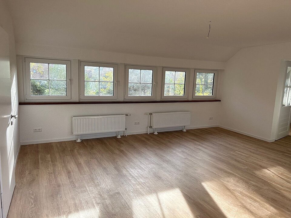 Leerer, sonnendurchfluteter Raum mit großen Fenstern und zwei Heizkörpern unter weißer Wand und hellem Holzfußboden.