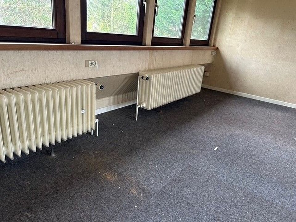 Altheizkörper unter einem Fenster an einer hellbeige Wand in einem leeren Raum mit dunklem Boden.